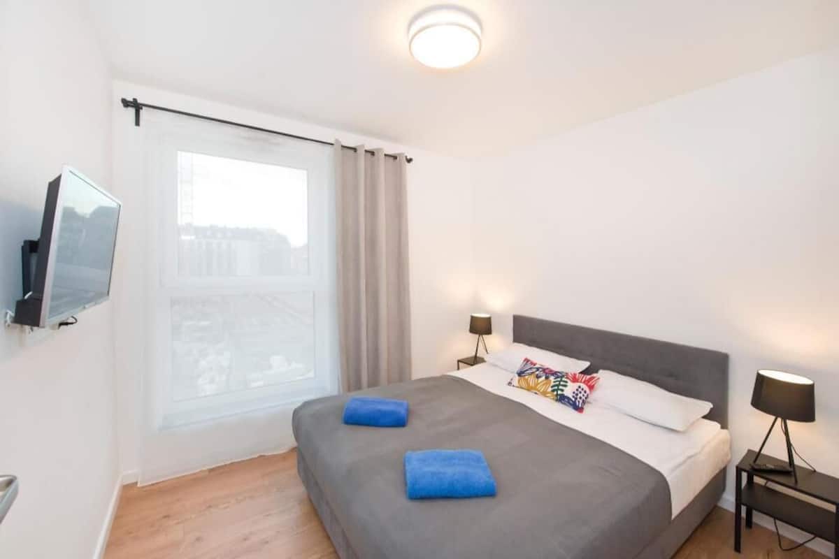 A hotel room with a bed, a TV mounted on the wall, a window with curtains, and a nightstand with a lamp.