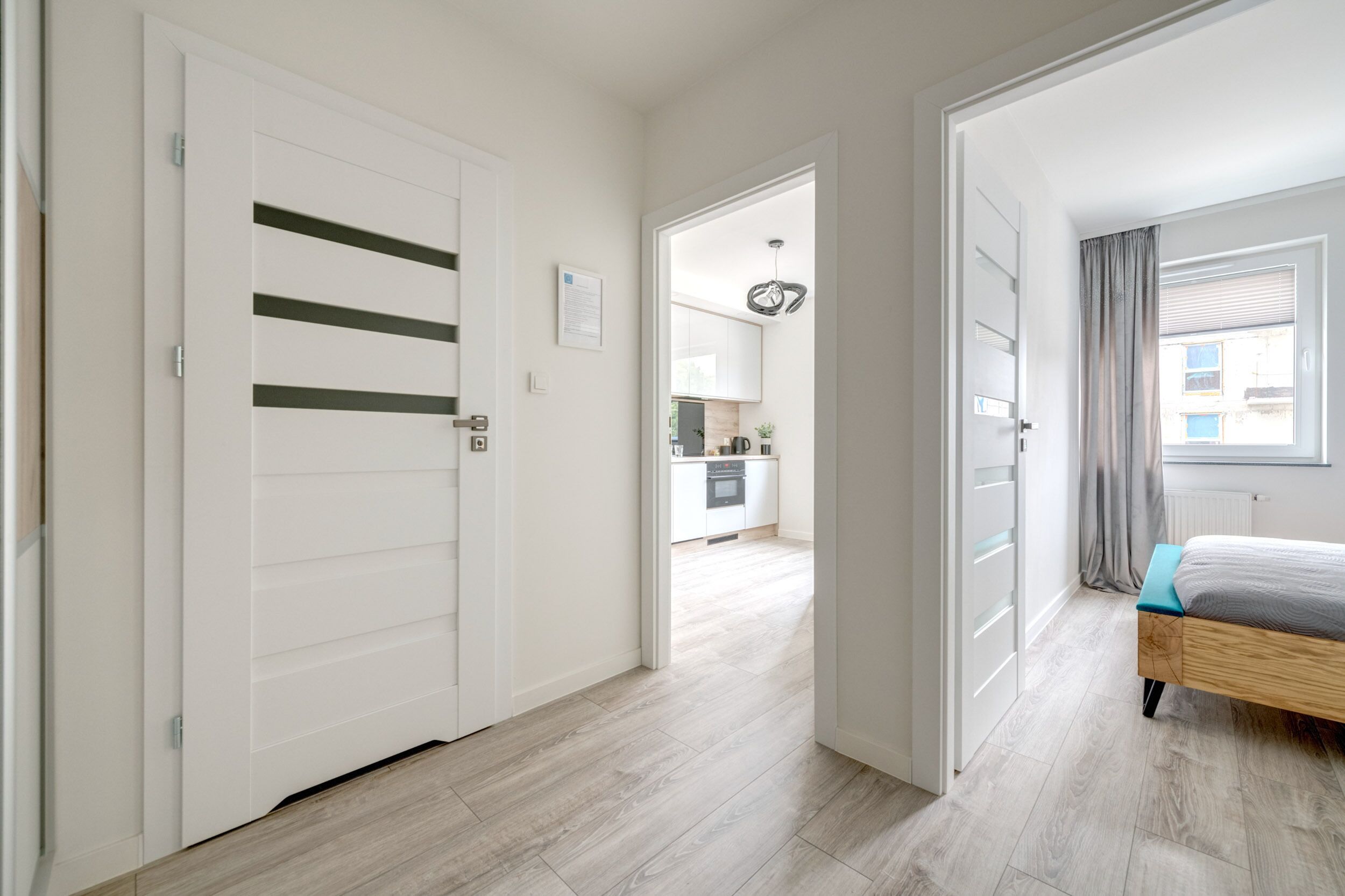 A modern bedroom with a wooden bed, a view to the kitchen, and a hallway leading to a door.