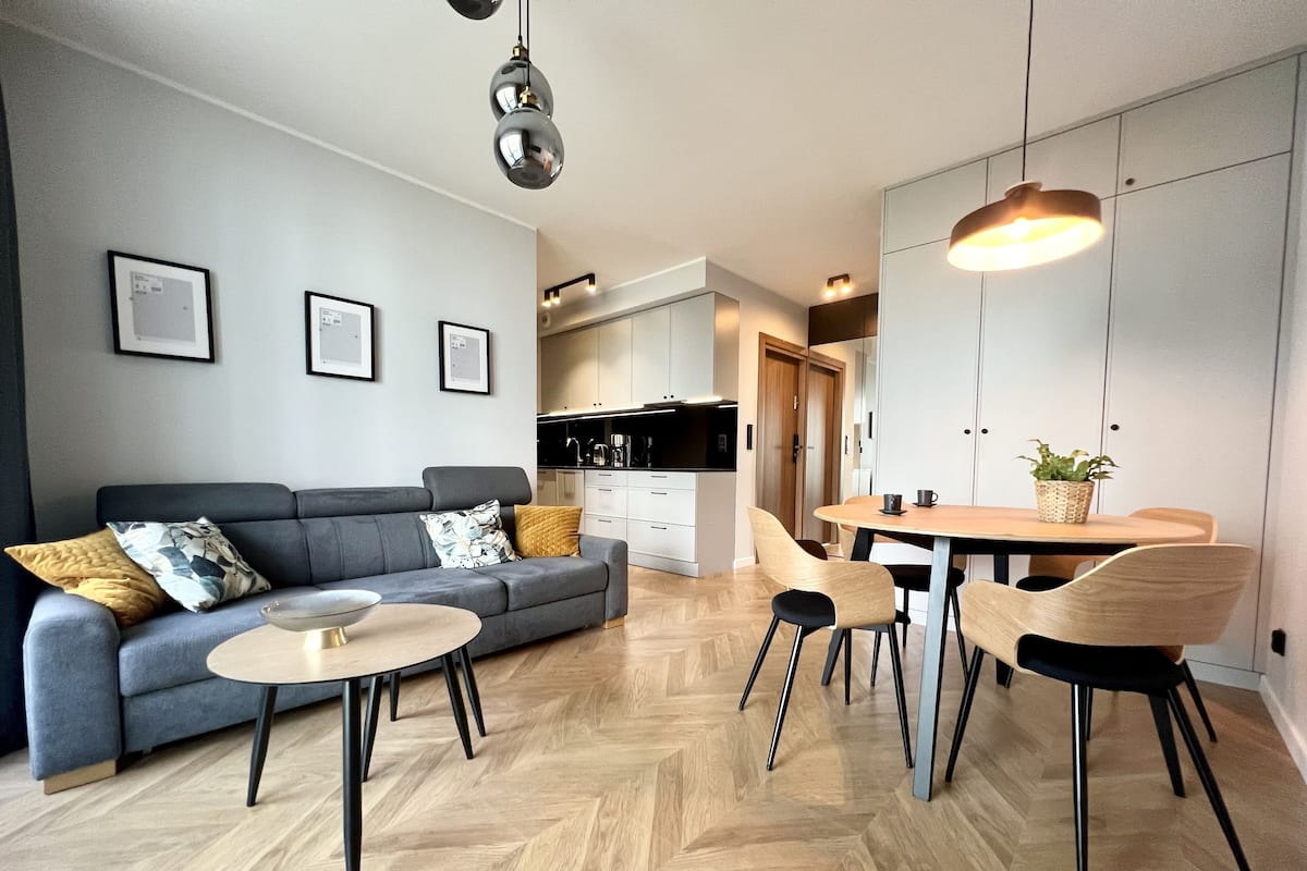 A modern living room with a grey sofa, a round coffee table, and a dining area with a wooden table and chairs. There is a kitchen in the background with white cabinets and a black countertop.