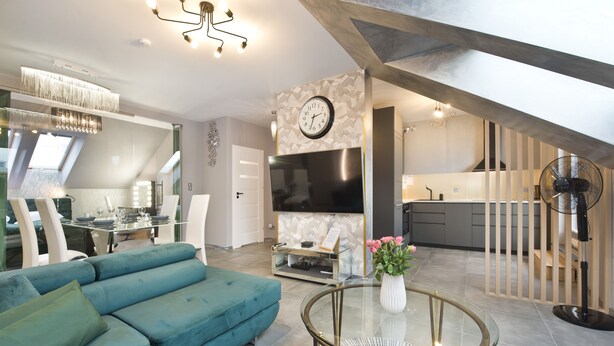 A modern living room with a teal sofa, a glass coffee table, and a kitchen area with a large TV and wooden cabinets. - Angielska Grobla by Q4 Apartments  (Gdansk)