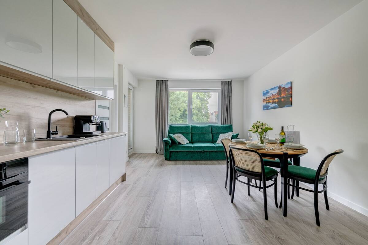 A modern kitchen with white cabinets and a wooden countertop, a green sofa in the living area, and a dining table set for four.