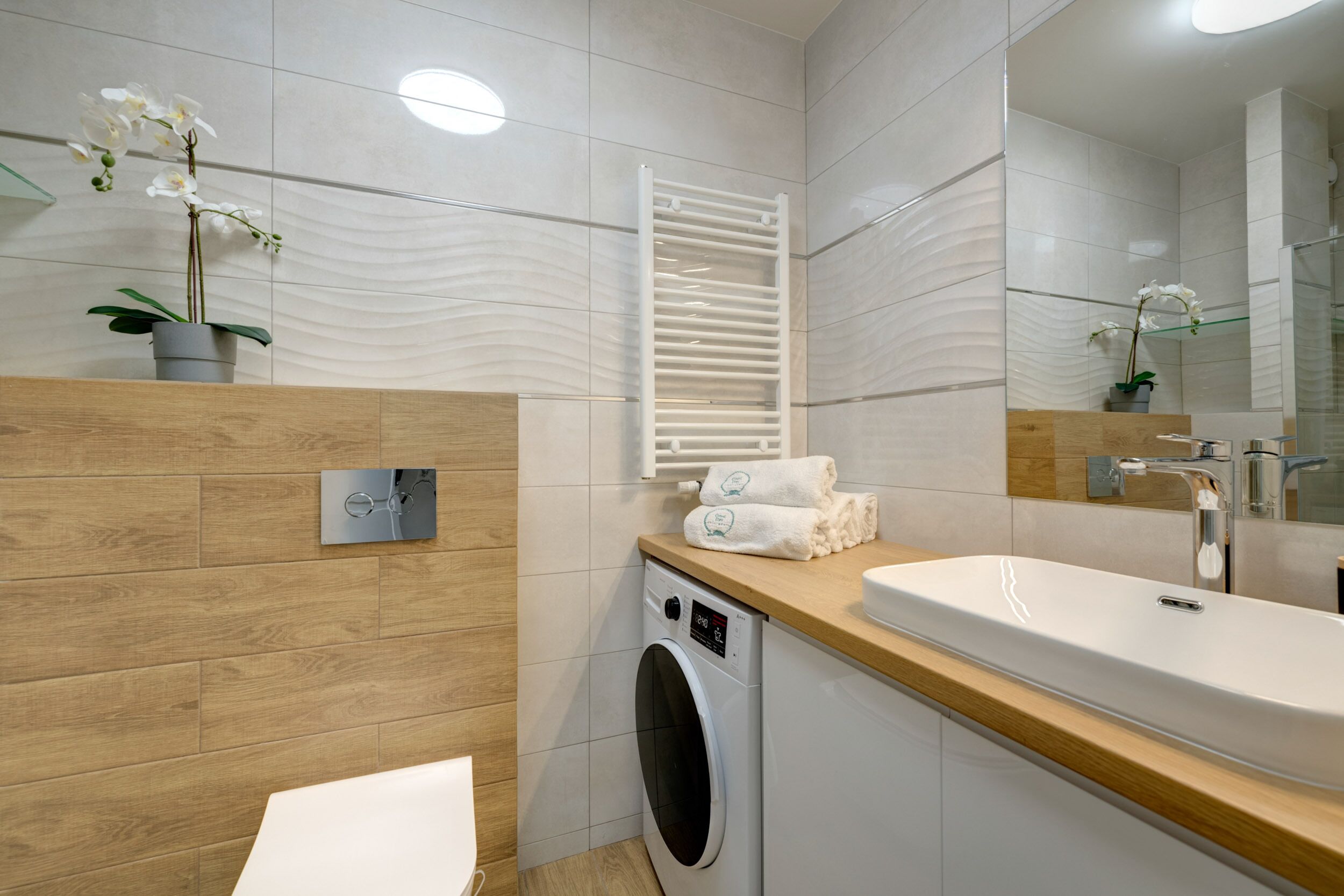 A modern bathroom with a wooden vanity, a built-in washing machine, a large mirror, and two potted orchids.