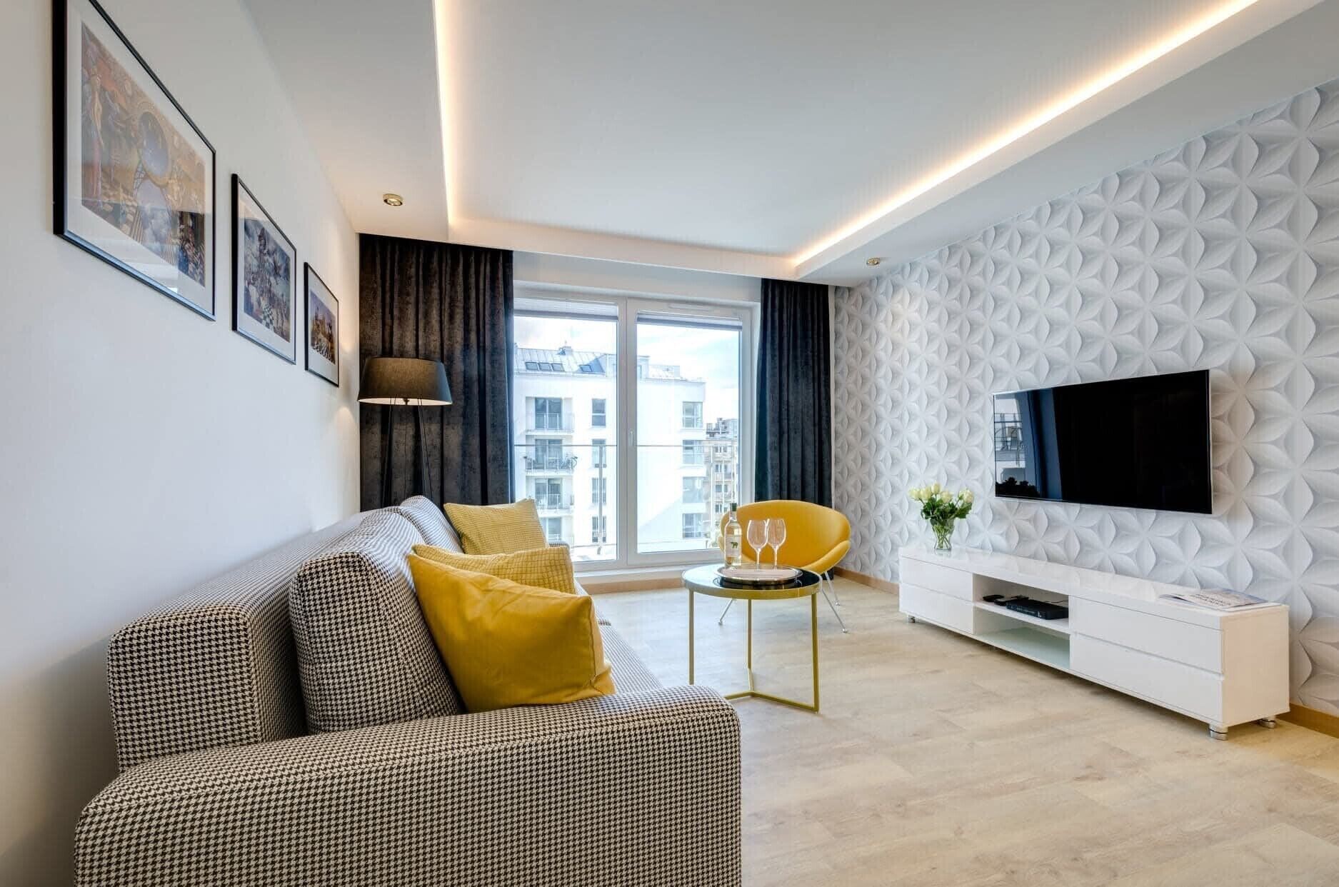 A modern living room with a houndstooth sofa, yellow cushions, a yellow chair, a white console table, and a flat-screen TV mounted on a textured wall.
