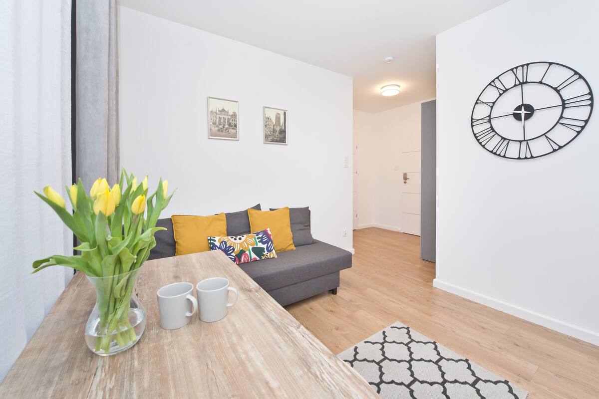 A modern living room with a grey sofa, a wooden coffee table with a vase of yellow tulips, two framed pictures on the wall, and a black clock.