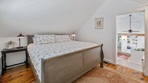 Un dormitorio con cama de madera, una mesita de noche con lámpara, edredón estampado con flores y vistas a una zona de cocina a través de una puerta abierta.