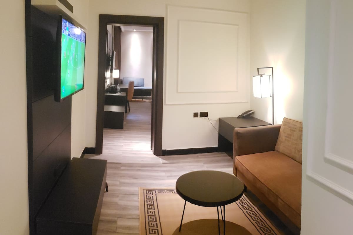 A hotel room with a flat-screen TV mounted on the wall, a sofa, a small round table, and a patterned rug.