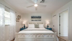 Une chambre à coucher avec un lit blanc, deux tables de chevet, un ventilateur de plafond et un tableau accroché au mur.
