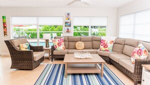 Un salon spacieux comprenant un canapé d’angle, deux fauteuils, une table basse et un ventilateur de plafond. De grandes fenêtres offrent une vue sur la verdure environnante.