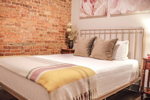 A bed with a white comforter and pillows, a brick wall, and a floral painting on the wall.