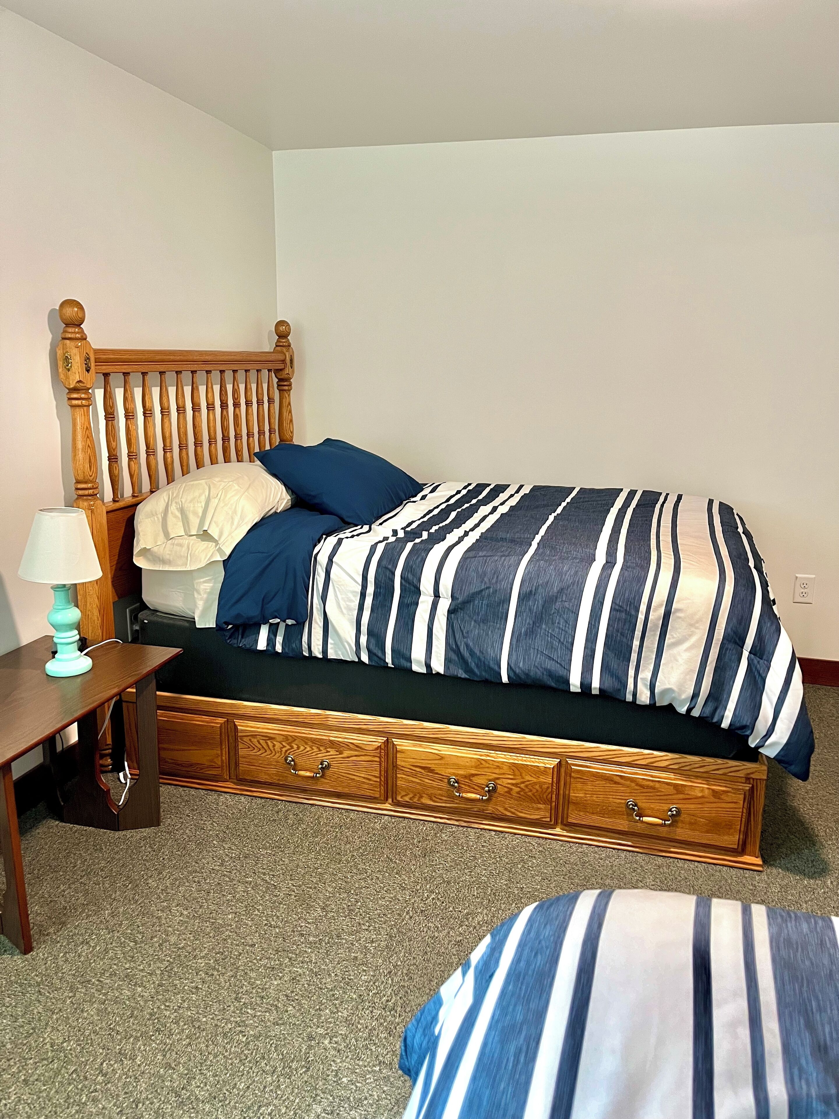 A bedroom with a wooden bed frame, a striped bedspread, a nightstand with a lamp, and a wooden dresser with drawers.