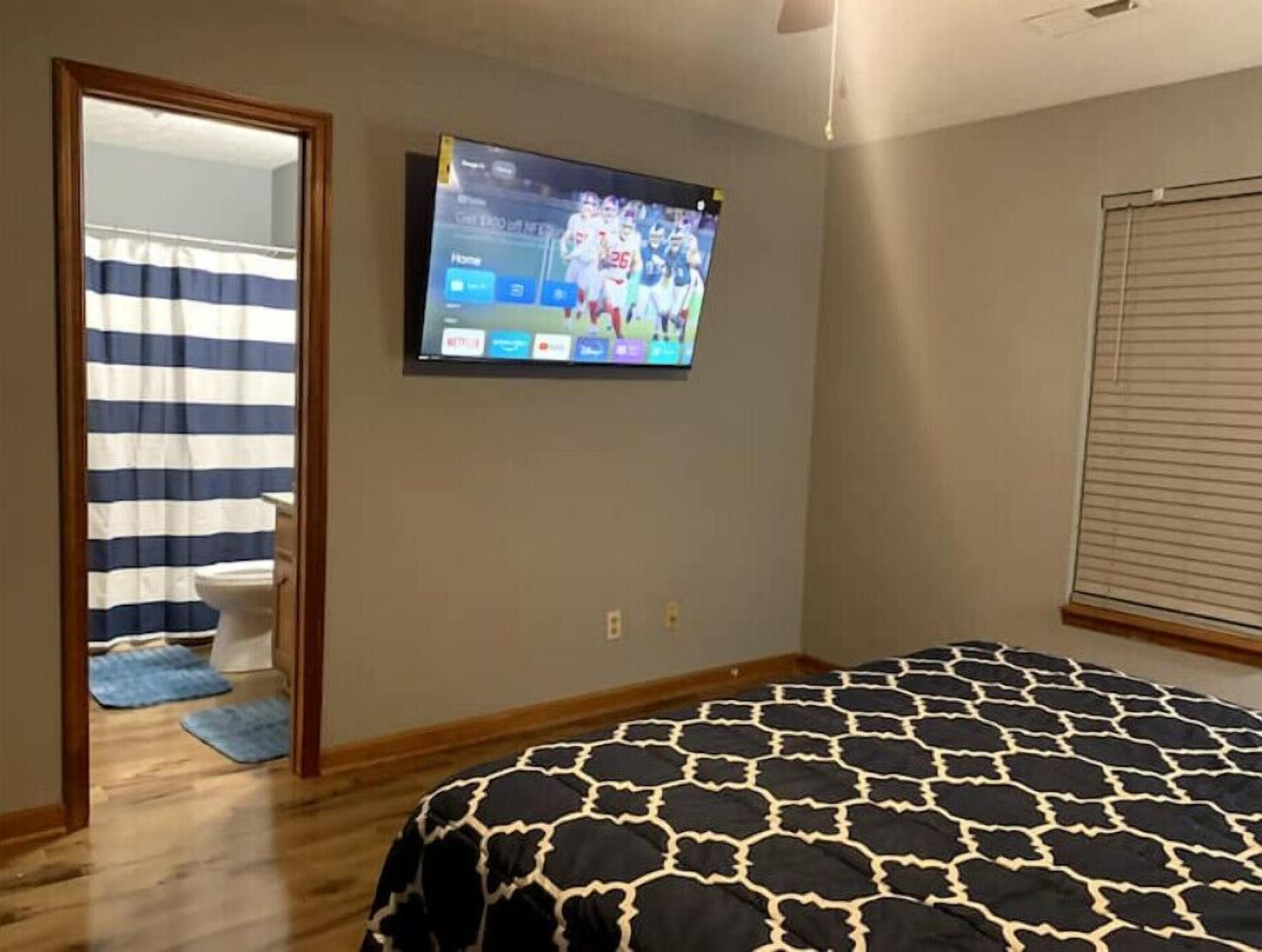 Ein Schlafzimmer mit Bett, Wandfernseher, Fenster mit Jalousien und einer Tür zu einem anderen Raum.