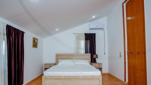 A bedroom with a wooden bed, two bedside tables, a nightstand, a window with curtains, an air conditioning unit, and a wooden door.