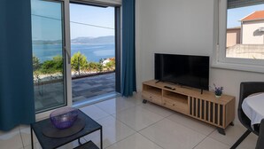 A modern living room with a large TV on a wooden stand, a glass table with a purple bowl, and a view of the sea through a sliding glass door.