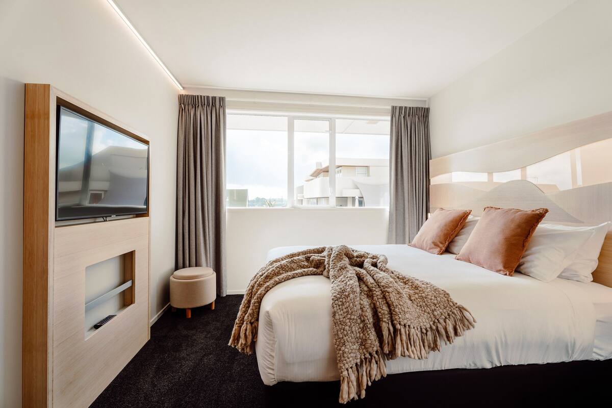 Een moderne hotelkamer met een groot bed, een flatscreen televisie aan de muur, een raam met gordijnen en uitzicht op de stad.