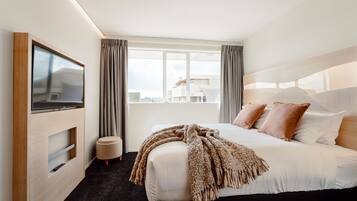 A modern hotel room with a large bed, a flat-screen TV mounted on the wall, a window with curtains, and a view of the cityscape.