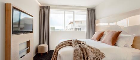 Ein modernes Hotelzimmer mit einem großen Bett, einem Wand-Fernseher, einem Fenster mit Vorhängen und Blick auf die Stadtlandschaft.