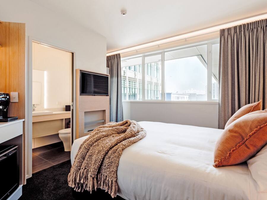 Habitación de hotel con cama, escritorio, cafetera, televisor y ventana con cortinas.