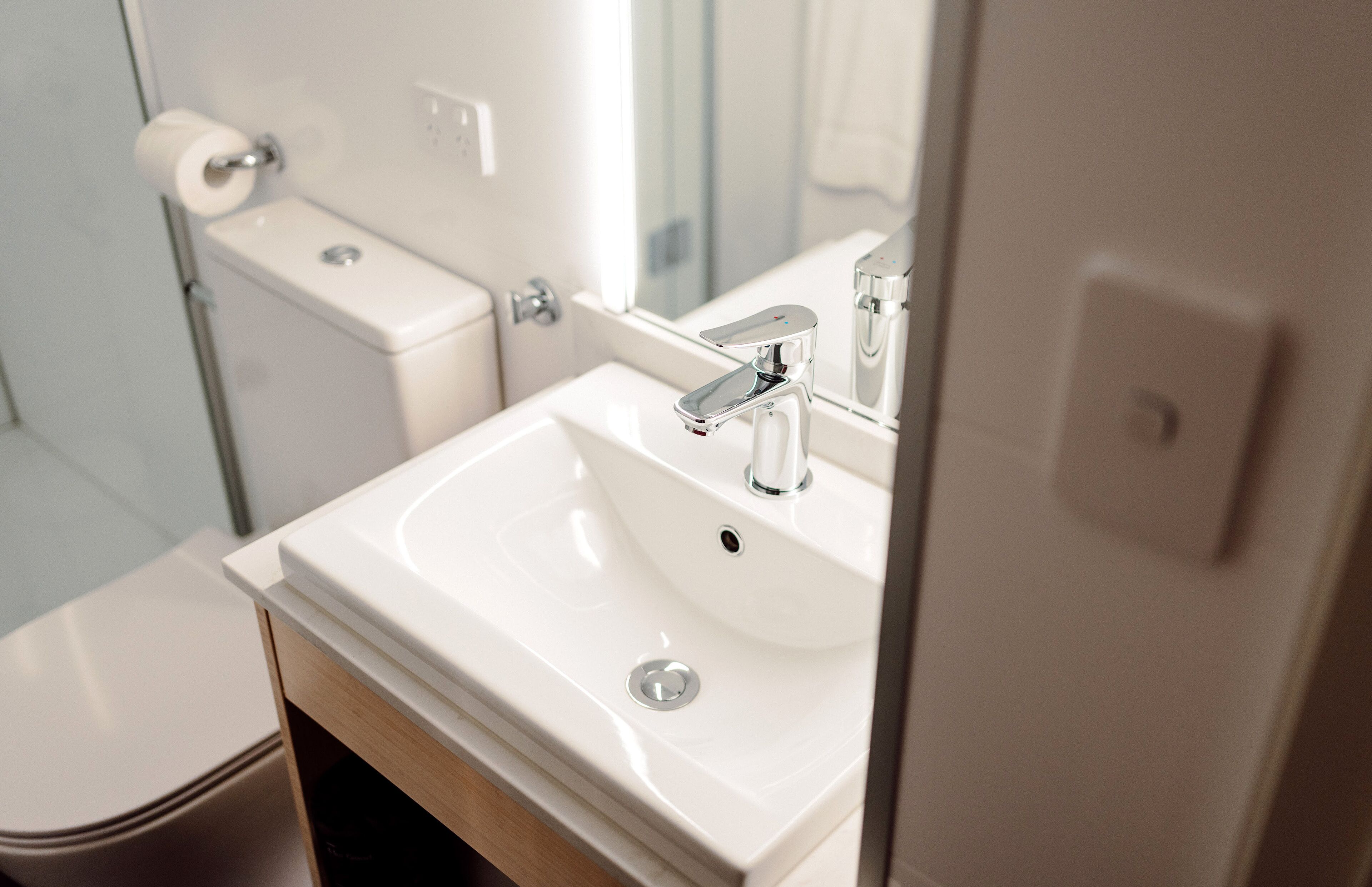 A modern bathroom with a white sink, chrome faucet, and a toilet with a paper towel holder.