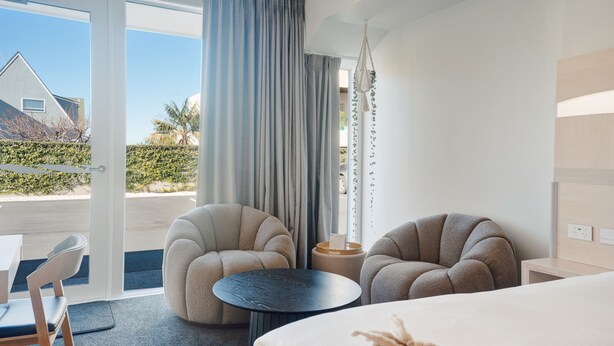 Een moderne hotelkamer met een groot bed, twee fauteuils, een tafeltje en uitzicht op het omliggende landschap.