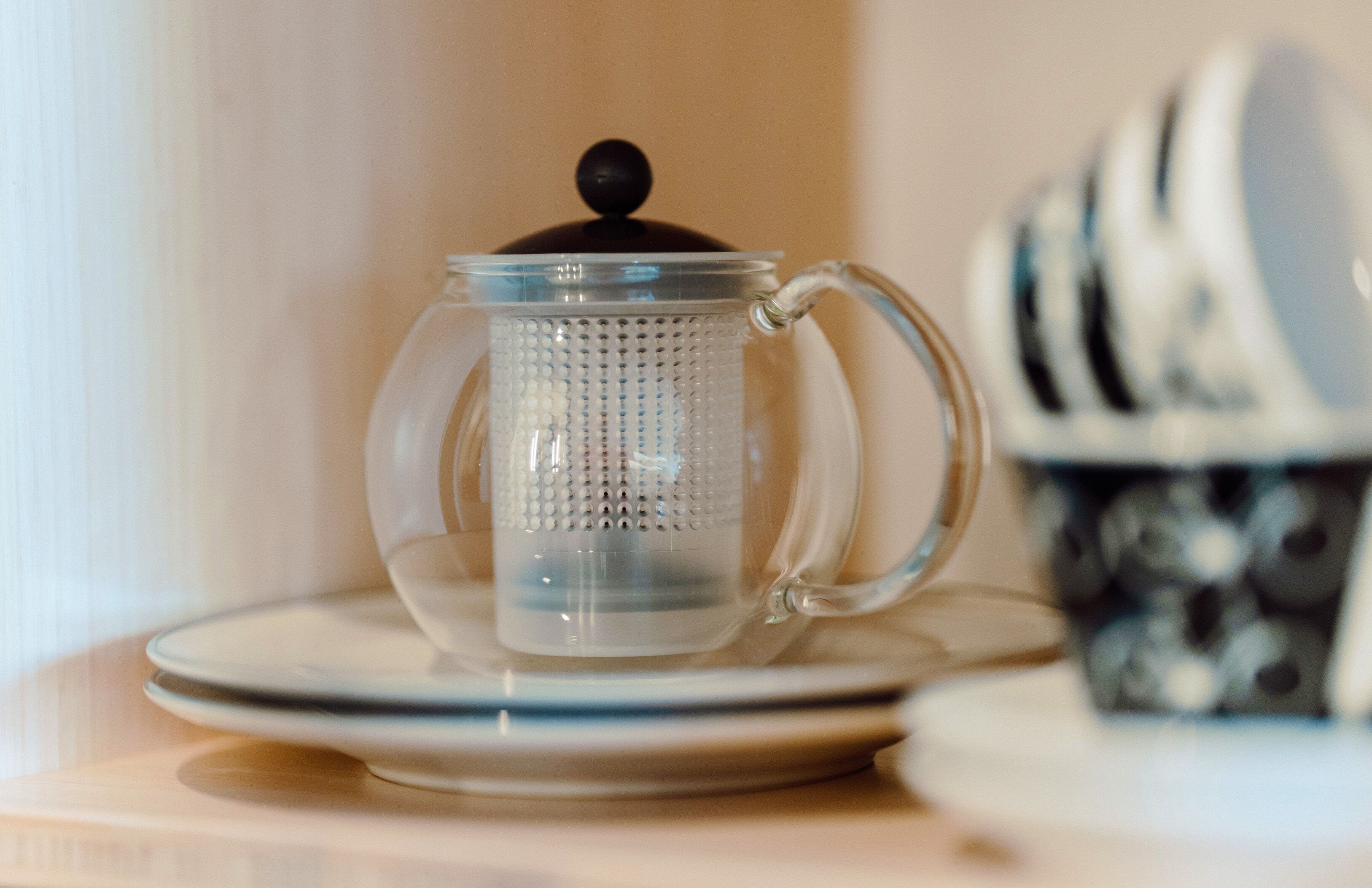 A clear glass teapot with a lid on a saucer, placed on a table with a stack of plates in the background.