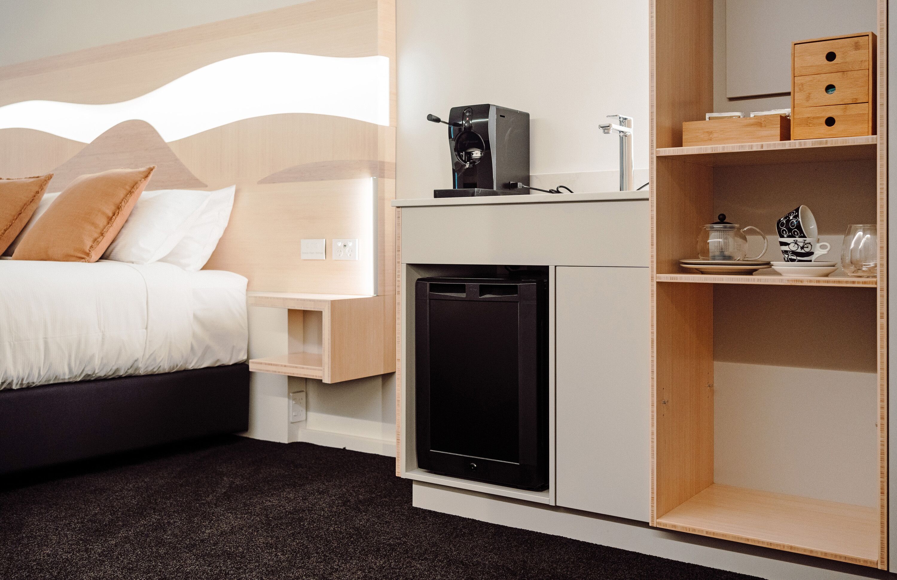 A modern hotel room with a bed, a coffee machine, a mini-fridge, and a shelf with tea and cups.