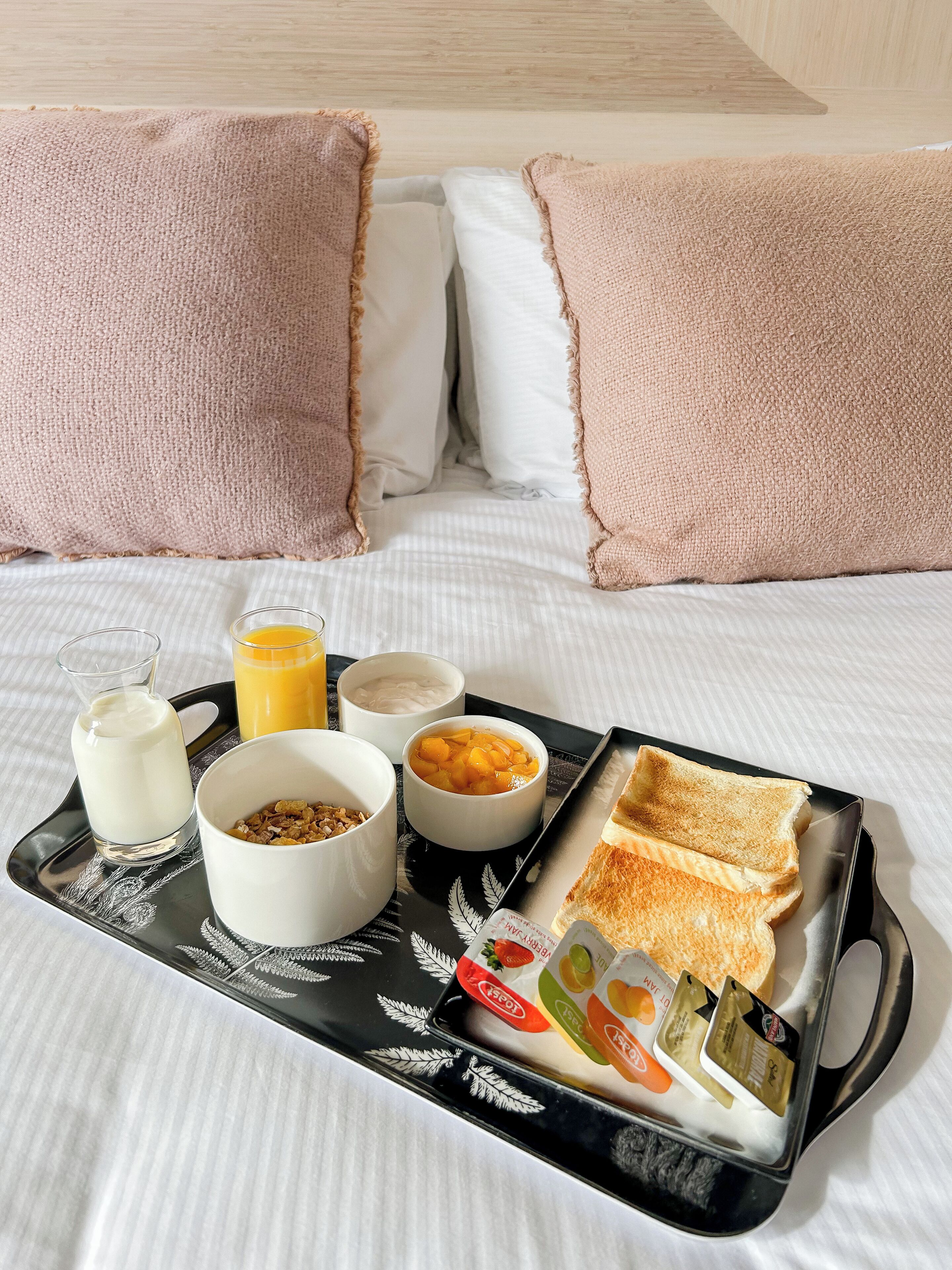 A neatly made breakfast tray with toast, cereal, milk, and juice on a bed with pink pillows.
