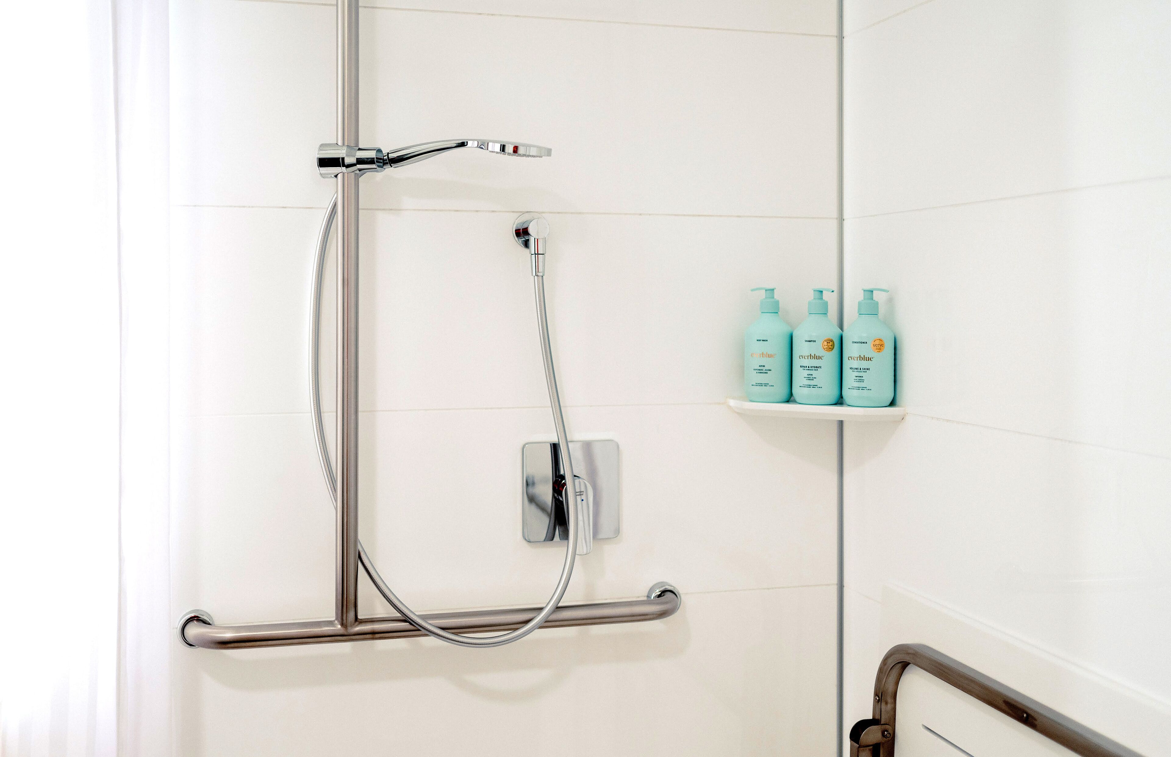 A shower with a handheld showerhead, a handheld shower attachment, and a grab bar. There are three bottles of Everblue body wash on a shelf.