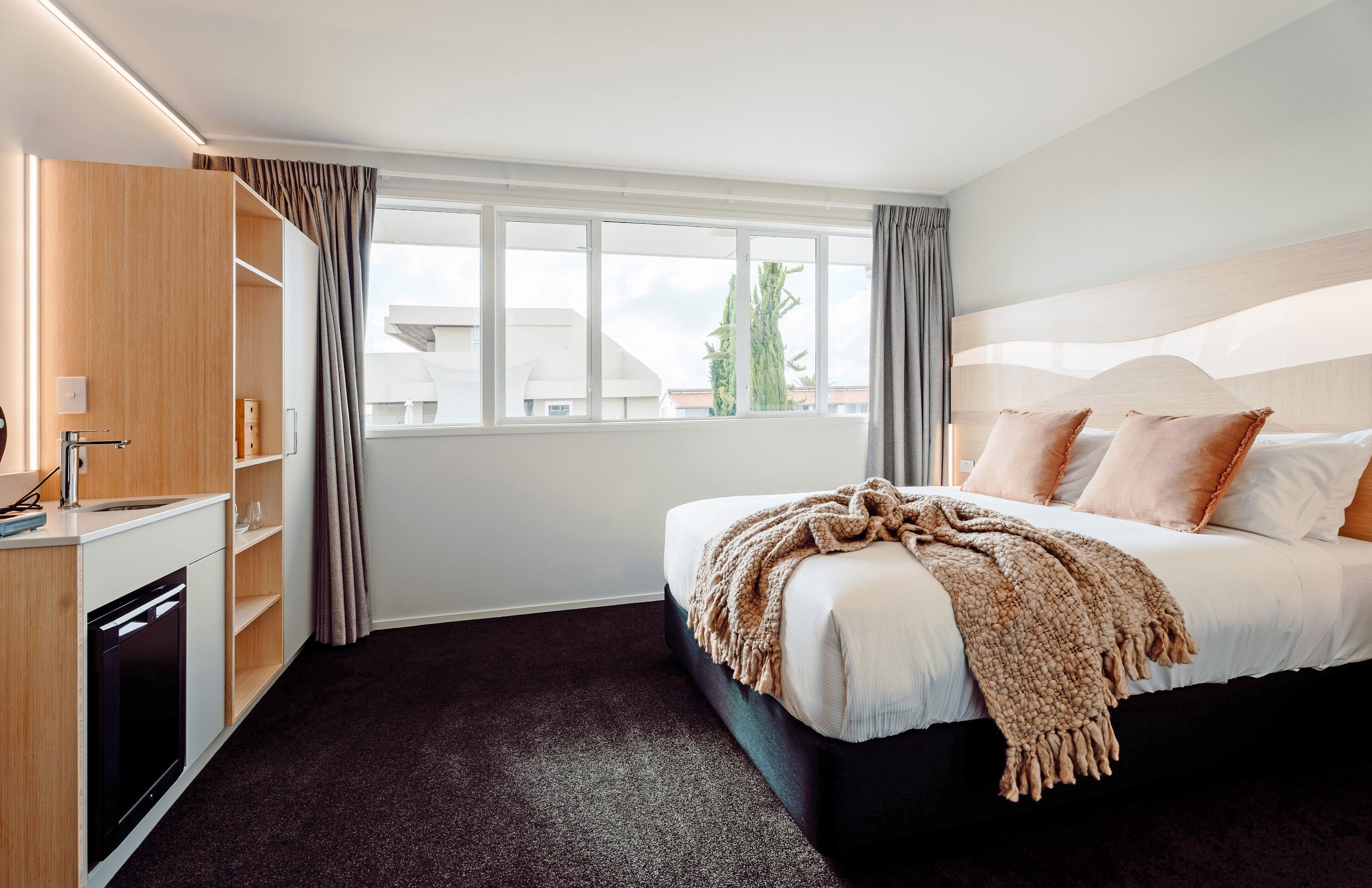 A hotel room with a bed, a built-in desk with a sink, and a window with curtains.