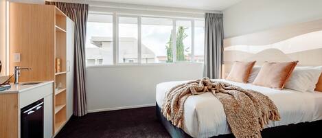 A hotel room with a bed, a built-in desk with a sink, and a window with curtains.