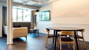 Een moderne hotelkamer met een zithoek, bestaande uit een ronde tafel met vier stoelen, een bank, een kleine bijzettafel met een vaas en een groot raam met gordijnen.