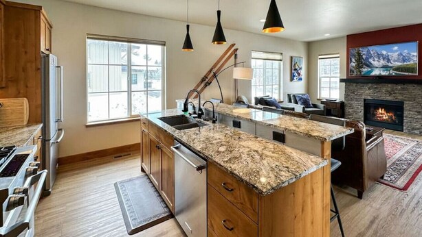 A modern kitchen with granite countertops, wooden cabinets, and a central island. There is a built-in oven, a sink, and a stove. The kitchen is adjacent to a living area with a fireplace and a large window. - Charming Tri-Level Condo in Downtown McCall (McCall)
