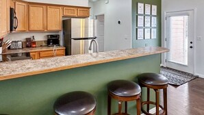 A kitchen with wooden cabinets, a granite countertop, a stainless steel refrigerator, and a bar stool area. - Relaxing Mountain Hideaway | Near Tamarack Resort (Donnelly)
