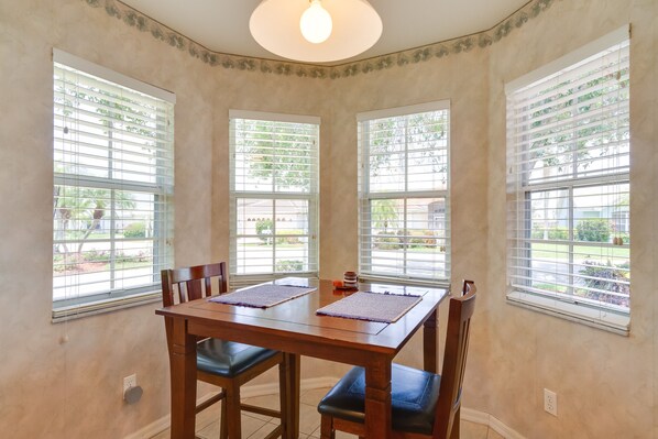 A dining area with a wooden table and chairs, three windows with blinds, and a ceiling light.