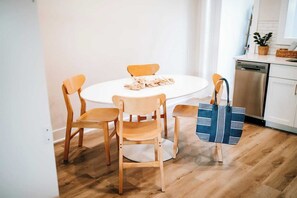 A dining area with a round table and four wooden chairs, a blue patterned bag on the floor, and a kitchen area with a dishwasher and cabinets.