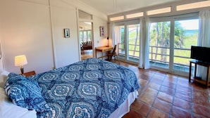 A bedroom with a large bed, a ceiling fan, a desk with a chair, a television, and large windows with curtains. - Heron Lane - Secluded, Waterfront, Incredible View (Cedar Key)