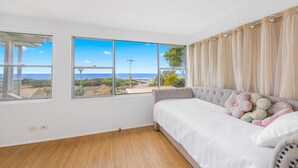 A bedroom with a large window offering a view of the sea and buildings, a grey sofa with floral cushions, and wooden flooring.