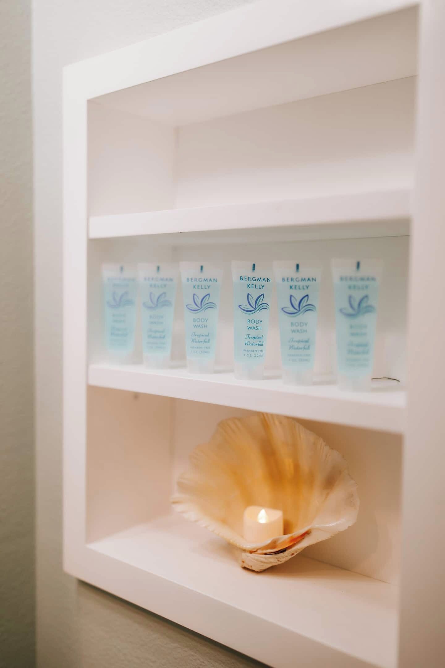 Une étagère blanche sur laquelle sont posées quatre bouteilles de gel douche Bergman Kelly et un porte-bougie en forme de coquillage.