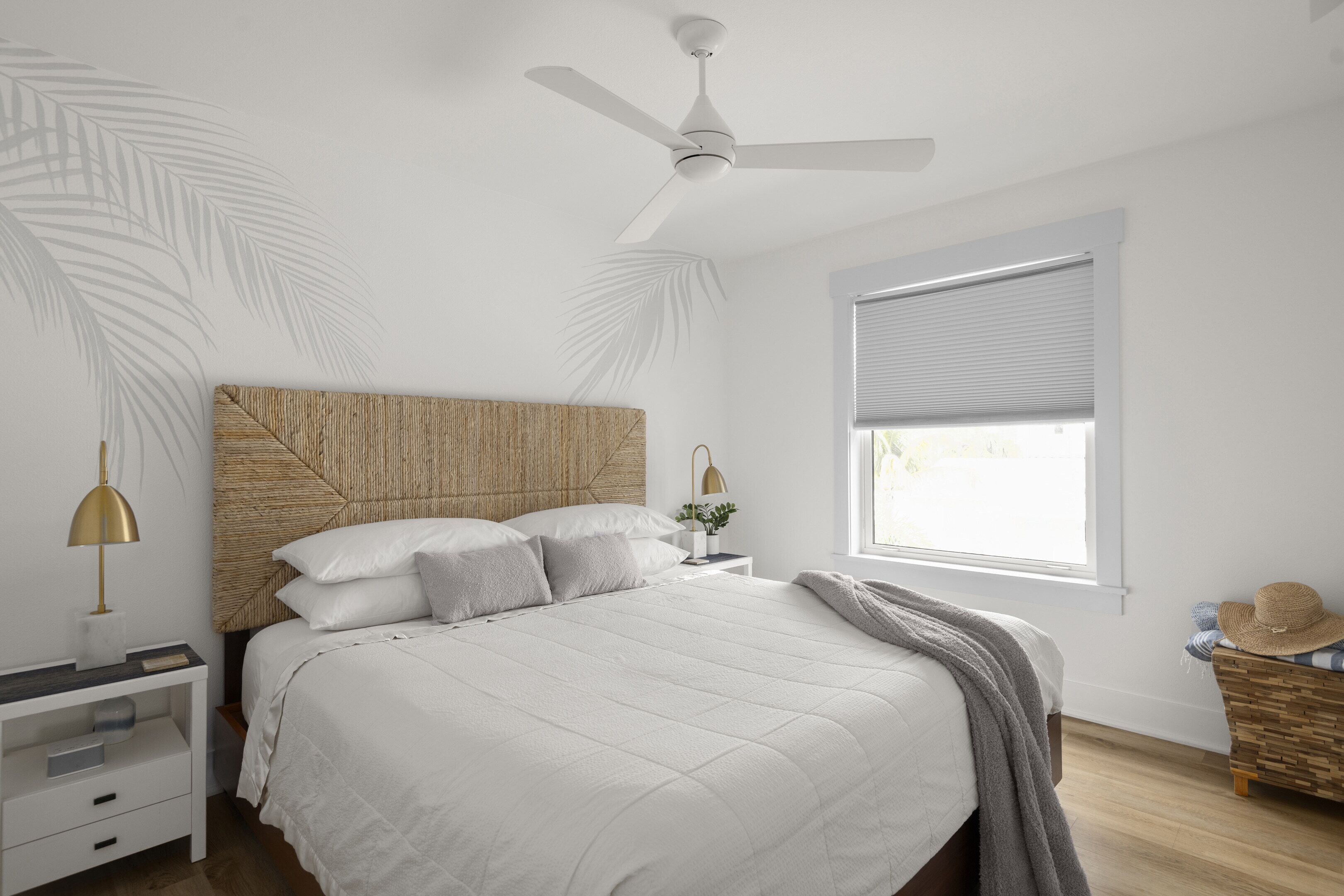 Un dormitorio con cama, mesita de noche, lámpara, ventilador de techo, ventana con persianas y una mesita auxiliar de madera con un sombrero.