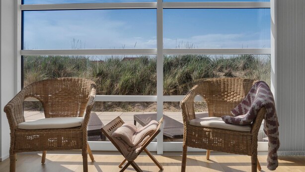 Ein Zimmer mit zwei Korbstühlen, die auf ein großes Fenster mit Blick auf einen Strand und hohes Gras zeigen.