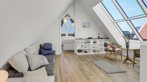 Ein Loft-Zimmer mit schrägen Wänden, einem Bett, einer Sitzecke mit Kissen, einem weißen Regal und einem großen Dachfenster.