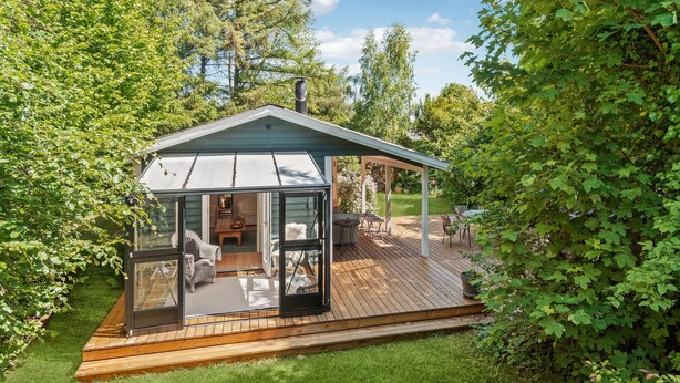 Een glazen terras met een houten terras, tuinmeubilair en uitzicht op het omliggende groen.