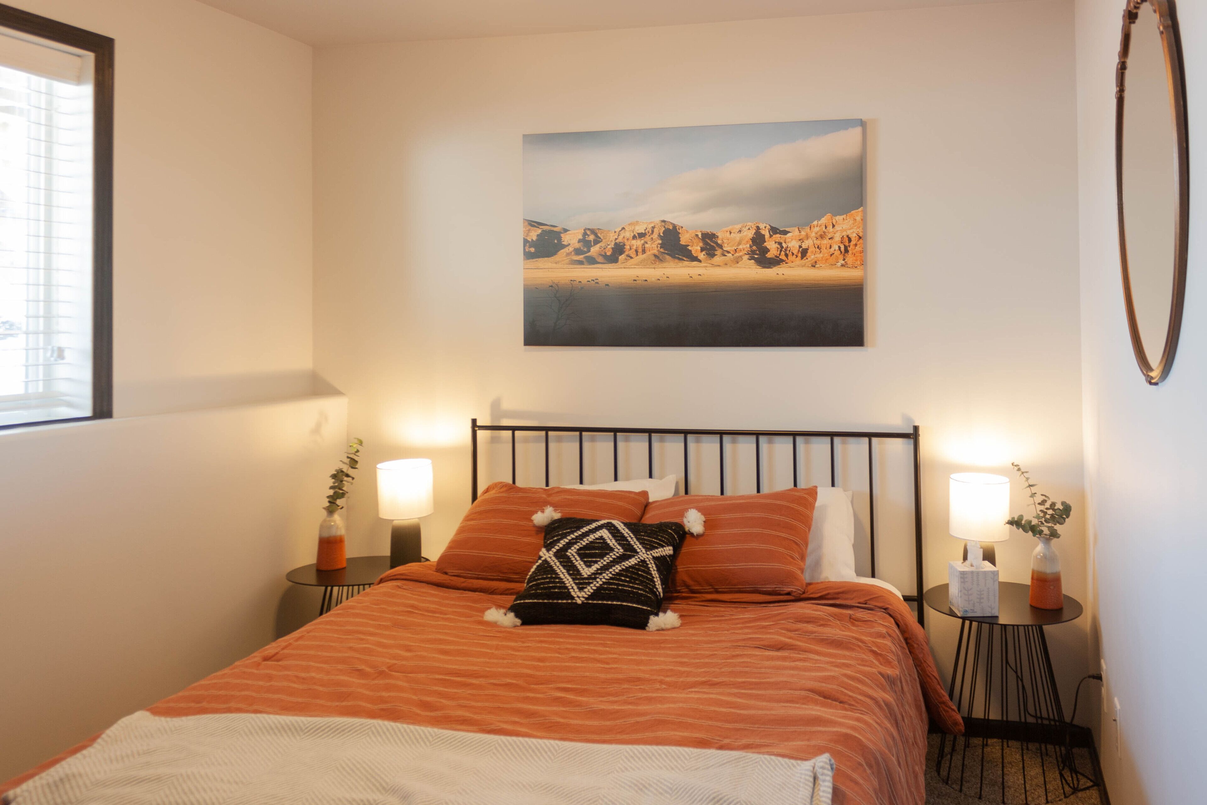 Una cama bien hecha con una colcha naranja, dos mesitas de noche con lámparas, un cuadro en la pared y una ventana con persianas.