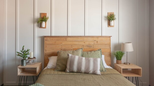 Un dormitorio con cabecera de madera, cama con ropa de cama verde, dos mesitas de noche con lámparas y plantas, y un estante montado en la pared con una planta.