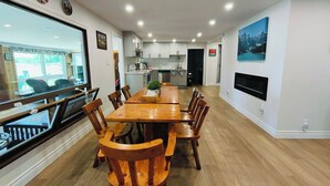 A spacious dining area with a long wooden table and chairs, a kitchen in the background, and a large window with a view of the outdoors. - 全景湖景私密临水度假屋 (Kawartha Lakes)