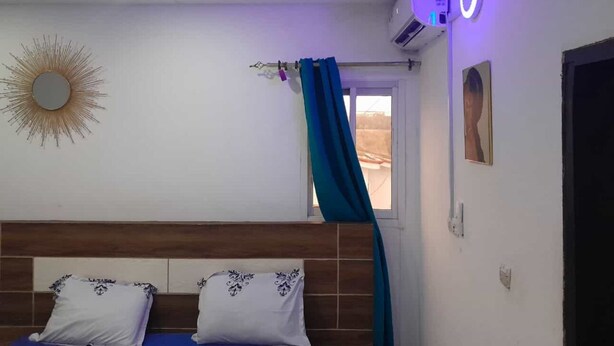 Une chambre à coucher avec une tête de lit en bois, une fenêtre avec des rideaux, un miroir fixé au mur et un appareil de climatisation.