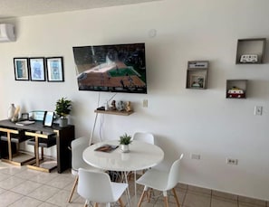 Un salon avec un téléviseur fixé au mur, une table de salle à manger ronde entourée de chaises blanches, une petite étagère avec des objets décoratifs et une table basse.