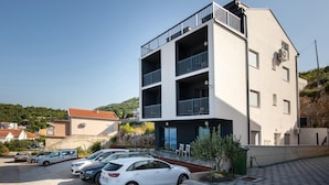 Un edificio de varios pisos con balcones, un estacionamiento con varios coches y un paisaje montañoso al fondo.