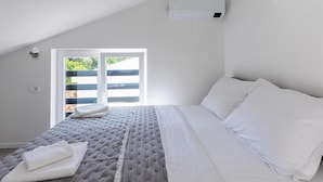 Un dormitorio con cama, ropa de cama blanca, una ventana con vistas a zonas verdes y un aire acondicionado instalado en el techo.
