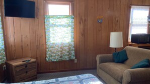 A room with wooden panel walls, a bed with a quilt, a sofa with cushions, a TV mounted on the wall, and a window with a curtain. - Cabin 2 - Reel and Trigger (Tom Township)