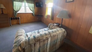 A room with wooden walls, a sofa, a small table, a lamp, and a window with curtains. - Cabin 3 - Reel and Trigger (Tom Township)